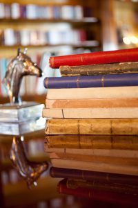 Antique books over a wooden luxury table. Defocused bookshelf on the back and a small metal horse head. Reflection on table.
